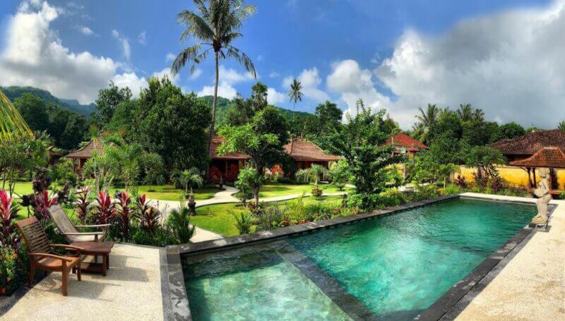 A landscaped tropical garden at Kubu Kangin Resort in Amed features a rectangular swimming pool, lounge chairs, lush greenery, and traditional-style villas under a partly cloudy blue sky.