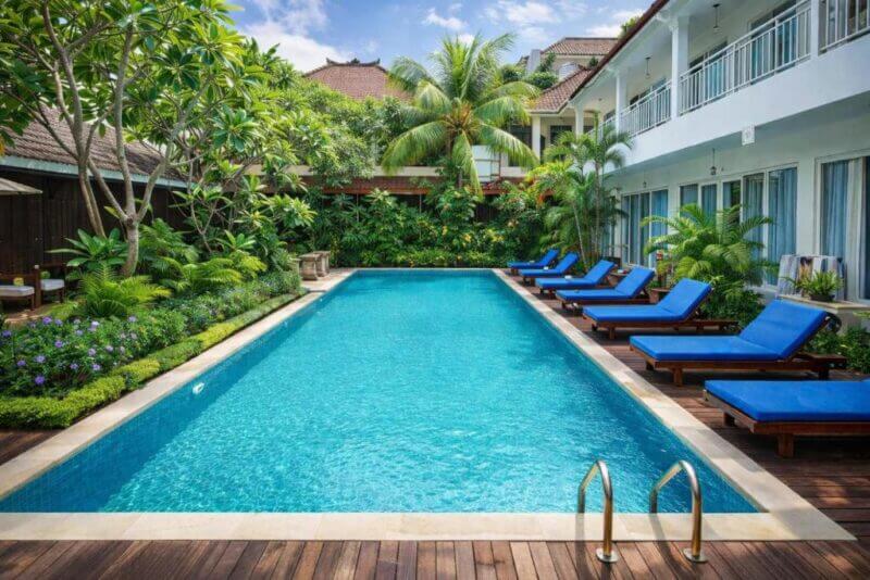 Rectangular outdoor swimming pool at Easy Surf Camp, Kuta, surrounded by blue lounge chairs, tropical plants, and a two-story building with balconies under a partly cloudy sky.