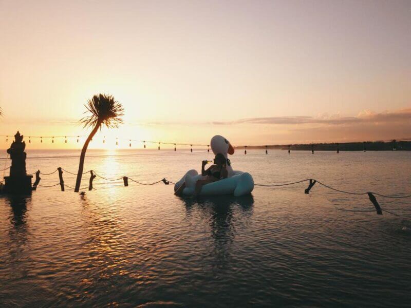 Person floats on an inflatable raft in a calm infinity pool at sunset at Nusa Veranda Ceningan, with a palm tree and decorative statue nearby, overlooking the ocean.