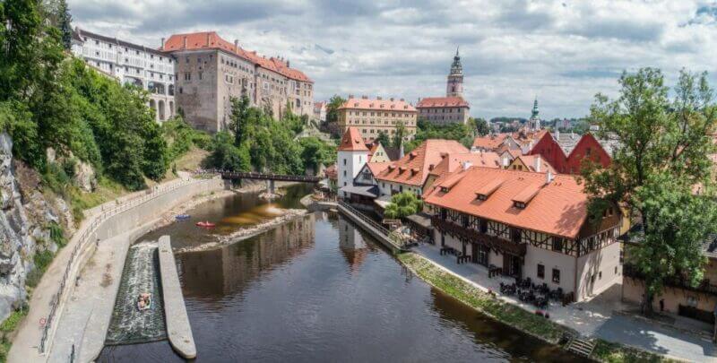 A view of Cesky Krumlov's river winding through its historic town with red-roofed buildings, a castle on a hill, and lush trees under a partly cloudy sky—perfectly seen from a cozy Garni hotel nearby.