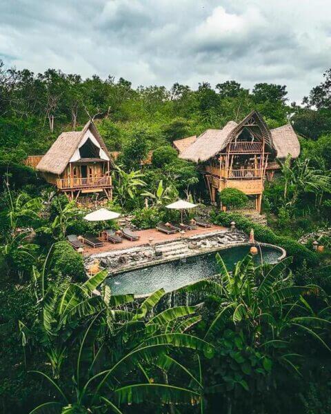 Three thatched-roof wooden villas at Green Suites Penida are surrounded by lush greenery, overlooking a stone-edged pool with lounge chairs and white umbrellas under a cloudy sky.