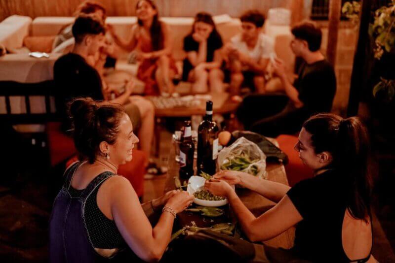 A group of backpackers sit around a table indoors in Berat, talking and sharing snacks, with drinks and food on the table. Two women in the foreground are smiling and eating together.