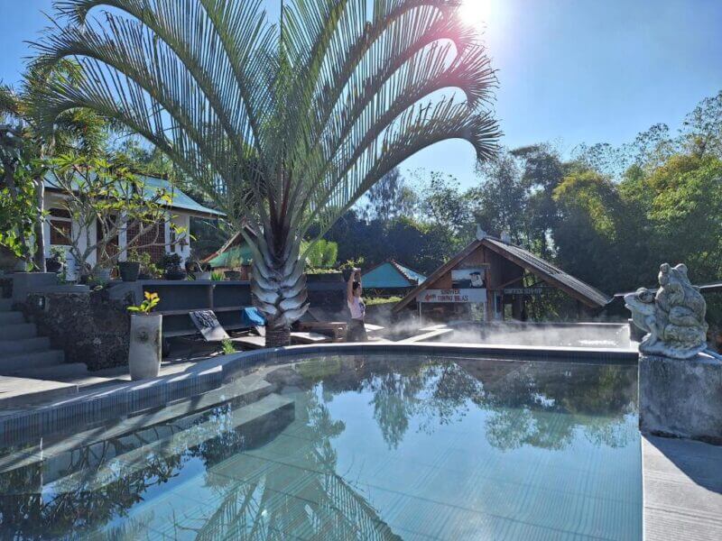 A person stands by a steaming outdoor pool surrounded by tropical plants and buildings, with the striking Black Lava Batur landscape visible under the clear blue sky and bright sunlight.