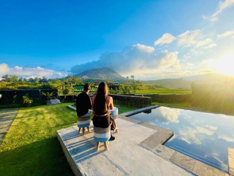 Two people sit by an outdoor pool facing the stunning Batur Panorama, with green fields, trees, and a mountain beneath a bright blue sky dotted with clouds.