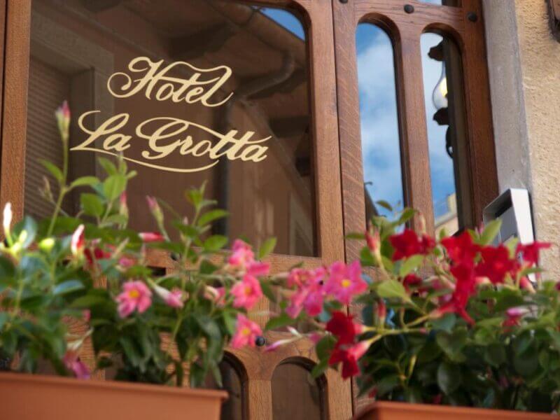 Wood-framed glass door with Hotel La Grotta written in gold lettering, partially obscured by potted pink and red flowers in the foreground, welcomes guests to this charming San Marino retreat.