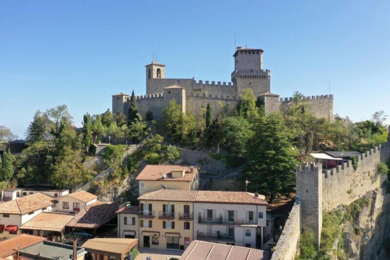 A large stone castle with towers sits on a hill above houses and trees in San Marino, surrounded by fortified walls under a clear blue sky, near the charming Hotel Rosa.