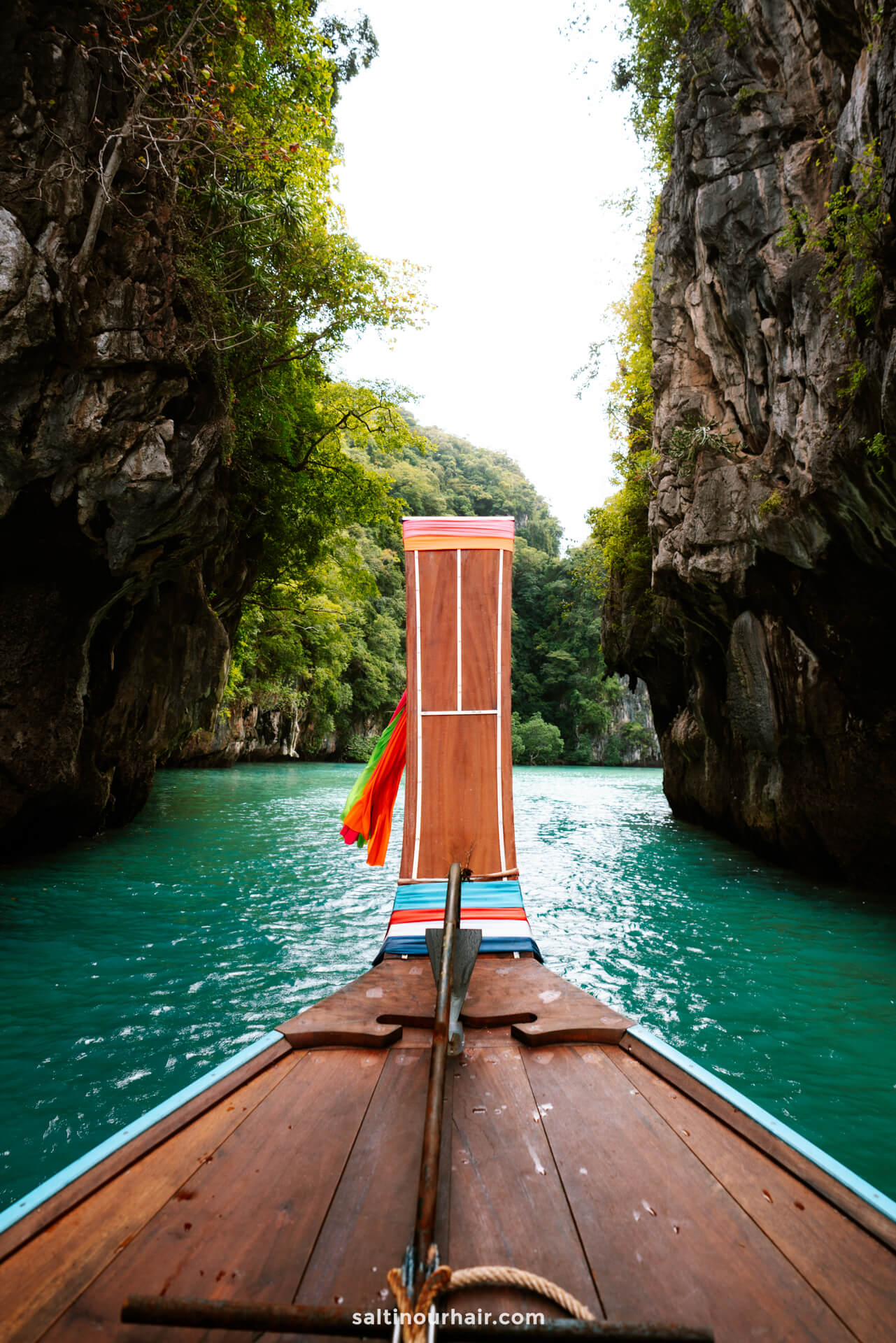 A wooden boat glides through turquoise water between two rocky cliffs covered in green vegetation, evoking the serene beauty often discovered after completing your Thailand arrival card.