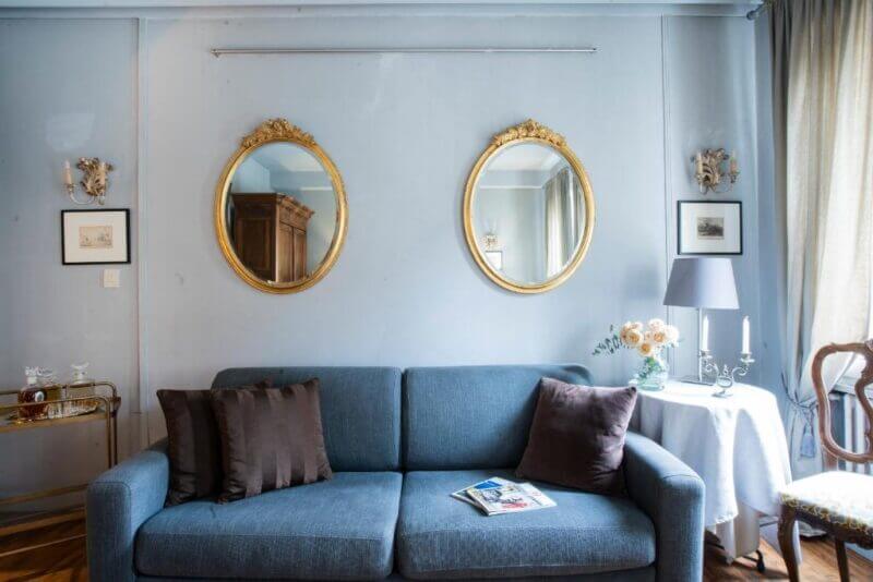 A blue Giò Venice sofa with brown cushions sits against a light blue wall with two oval gold-framed mirrors. A side table with flowers and lamp is on the right; magazines rest on the sofa.