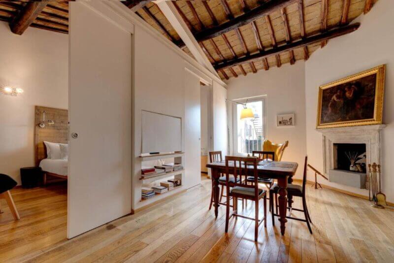 Open-plan living space in Casa Modelli, Rome, featuring wooden floors, exposed beam ceiling, dining table, fireplace, armchair, bookshelf, and a partially visible bedroom behind a sliding door.