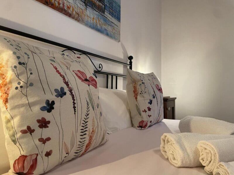 A neatly made bed with two floral patterned pillows, white sheets, and three rolled white towels placed on top at La casa di nonna in Montemerano, with a painting hanging above the headboard.