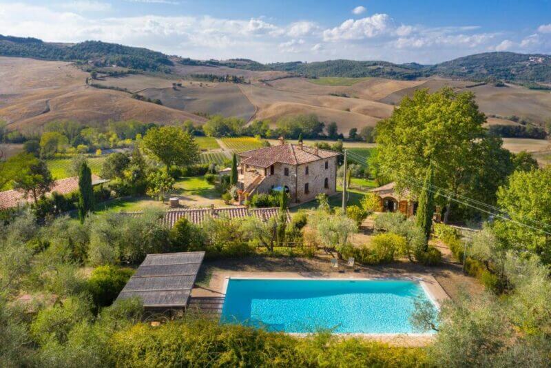 Aerial view of a rustic stone house with a swimming pool, nestled amid trees and hills in the Montepulciano countryside, capturing the charm of an Agriturismo under a partly cloudy sky.