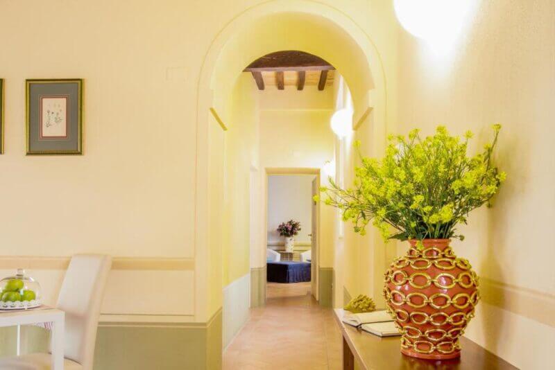 A hallway with light yellow walls, decorative vase with yellow flowers on a table, and framed pictures on the wall; at the end of the hall, a room inspired by La Chiocarella Pienza reveals a bouquet in warm sunlight.