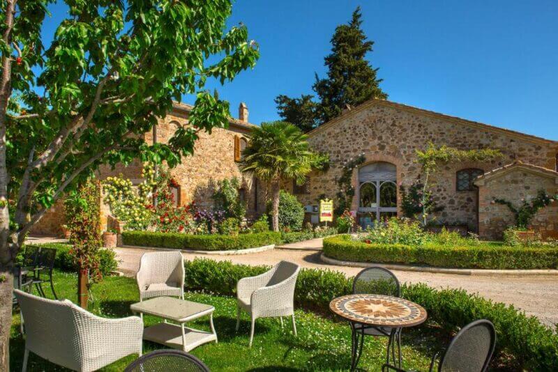 Stone buildings with arched windows and lush gardens surround outdoor seating areas with tables and chairs under a clear blue sky at Castello di Spedaletto, near the charming town of Pienza in Tuscany.