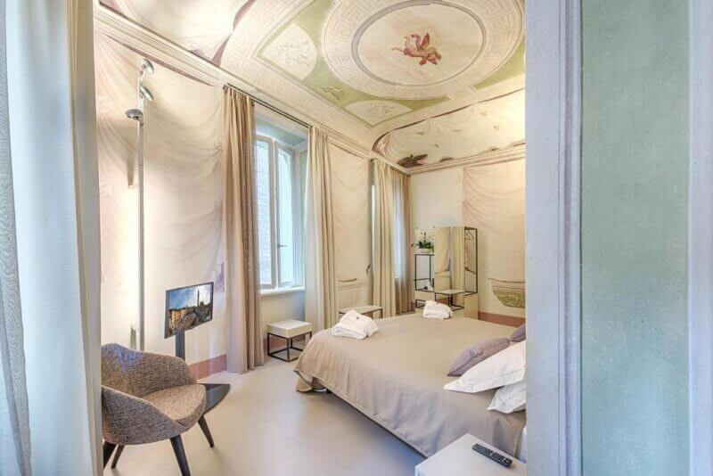 Elegant bedroom in a historic residence Siena with a frescoed ceiling, double bed, armchair, modern lighting, robes, large window, mirror, and muted pastel decor.