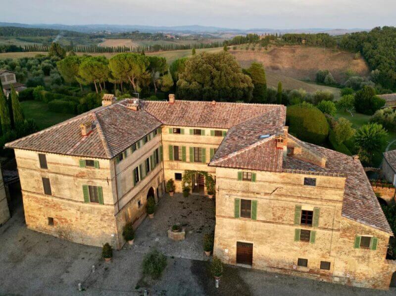 Aerial view of Ca' Bianca, a large, historic stone villa near Siena with tile roofing, surrounded by trees and rolling countryside.