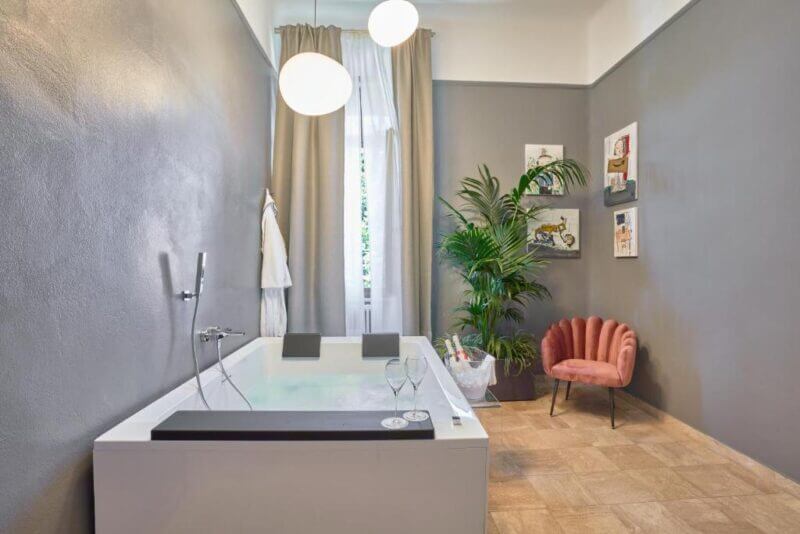Modern bathroom at Rif Hotel in Pisa, featuring a rectangular bathtub, two hanging lights, a pink velvet chair, potted plants, wall art, a robe, and light tan tile flooring for a chic hotel experience.