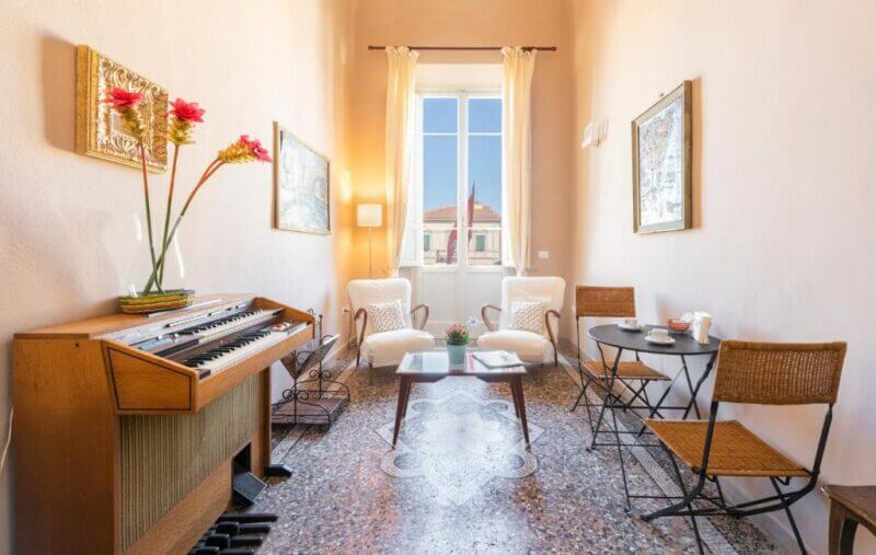 A bright living room with a keyboard, chairs, small tables, and artwork on the walls offers relaxing views reminiscent of Pisa’s charm, with sunlight streaming through sheer curtains like those in Torre Garbata.
