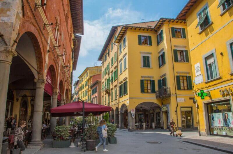 Pedestrian street in Rinascimento Pisa with yellow and red buildings, arched walkways, outdoor café seating, and people strolling on a sunny day.
