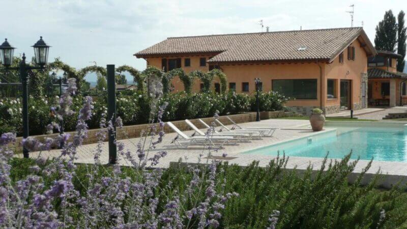 A modern house with a tiled roof stands behind a pool at Il Fienile di Assisi, surrounded by lounge chairs, plants, and flowering shrubs in an inviting outdoor setting.