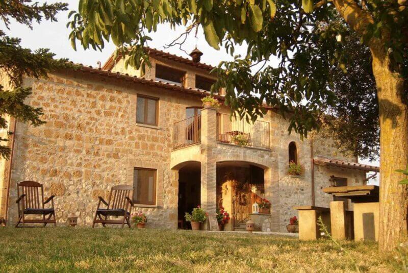 A rustic stone house with a tiled roof, arched entryway, and small balcony, this charming agriturismo near Orvieto is surrounded by trees and lawn, featuring two wooden chairs in the yard for relaxing afternoons.