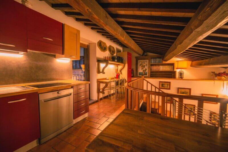 Loft-style kitchen and dining area with exposed wooden beams, red cabinets, tiled floor, and a staircase leading to a lower level. Warm lighting throughout the space makes it a cozy choice for Book and Bed Orvieto accommodation.