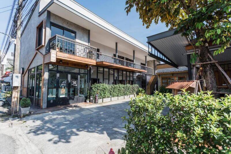 Modern two-story building with large windows, a balcony, and potted plants along the entrance, situated on a sunny day with trees nearby—this is Sleepy House Chiang Rai at its finest.