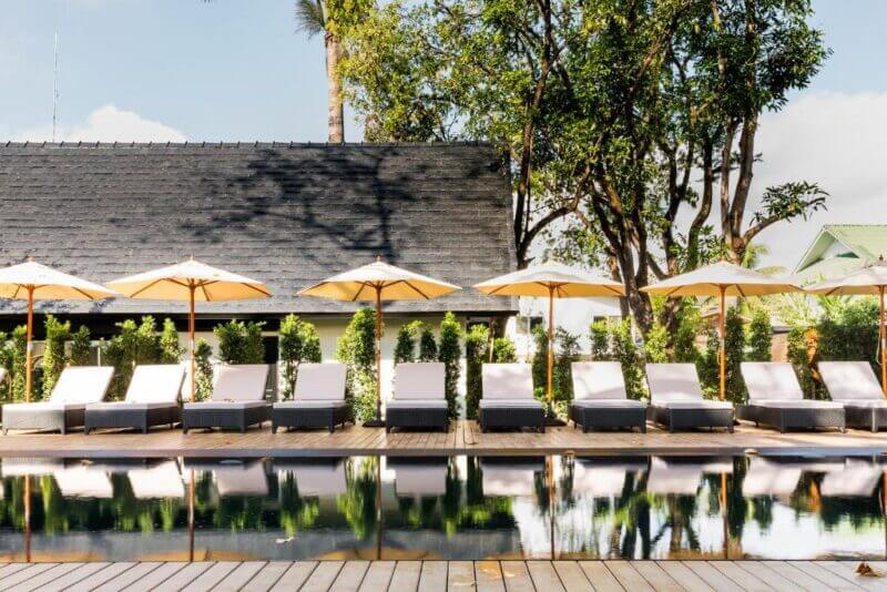 Row of white lounge chairs with beige umbrellas beside a reflective swimming pool at Mora Boutique, with trees and Chiang Rai buildings in the background under a clear sky.