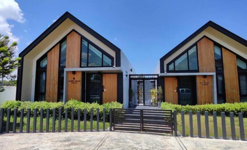 Two modern, A-frame villas with large front windows, wooden paneling, black trim, and a shared central gate under a clear blue sky at Baan Ga Cha Villa in Chiang Rai.