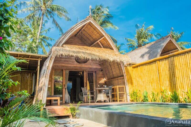 A tropical wooden villa at Anahata Gili Air with a thatched roof, open patio, outdoor seating, small pool, and surrounded by palm trees and bamboo fencing—a peaceful Gili Air accommodation under a clear blue sky.