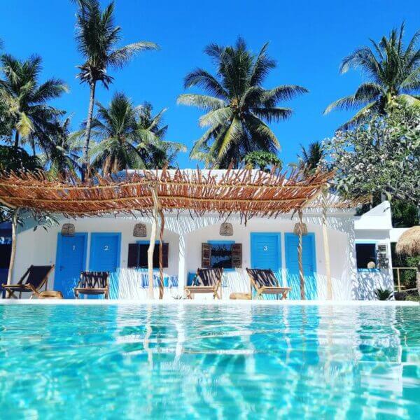 A small white building with blue doors sits behind a clear swimming pool at the Koho Air Hotel, surrounded by palm trees under a bright blue sky on Gili Air.