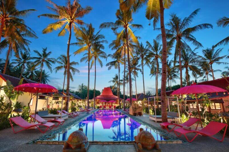 Outdoor pool at PinkCoco Gili Air, surrounded by palm trees, pink lounge chairs, and umbrellas at the resort during sunset with clear skies.
