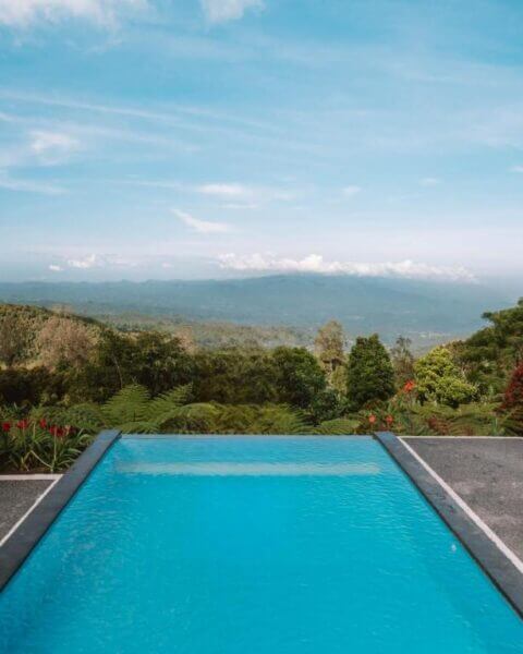 A rectangular swimming pool with clear blue water at Munduk Menir Villas overlooks a lush green landscape and distant mountains under a partly cloudy sky.