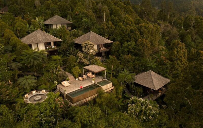 Aerial view of several wooden Munduk Cabins with thatched roofs surrounded by dense green forest, featuring a private pool and a circular hot tub.