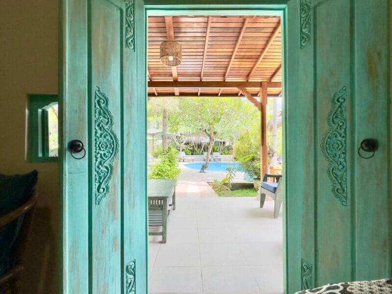 View through an open turquoise wooden door at Aashaya Jasri Resort, revealing a covered patio with a pool, lush greenery, and inviting outdoor seating in beautiful Bali.