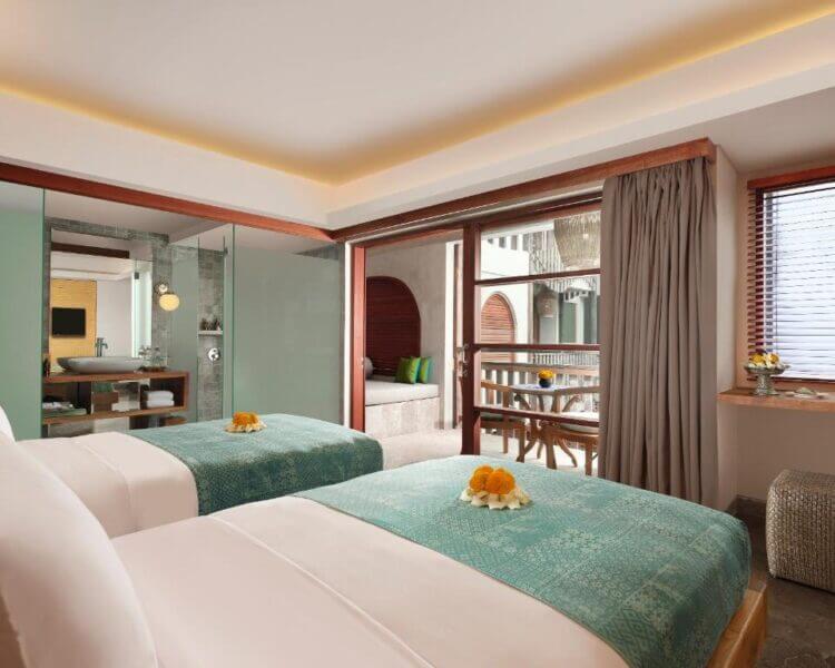 A modern Nusa Dua hotel room at Amnaya Resort features two twin beds with teal runners, a glass-walled bathroom, a balcony with seating, and large windows that fill the resort space with natural light.