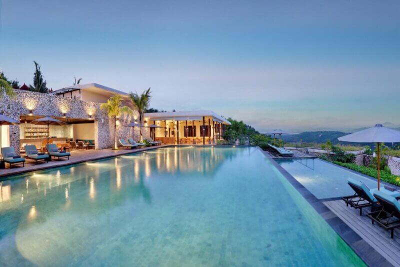Modern luxury villa with an infinity pool, outdoor lounge area, palm trees, and lounge chairs at MAUA Nusa Penida hotel, overlooking a scenic landscape at sunset.