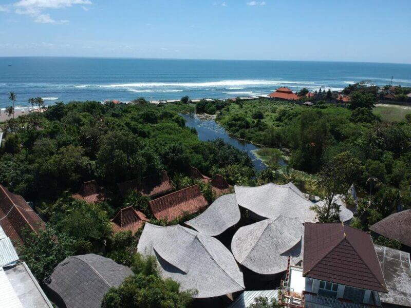 Aerial view of a coastal area with dense greenery, scattered buildings with sloped roofs, a small river, and the ocean in the background—an idyllic scene reminiscent of a Bali resort like ZIN Canggu Resort under a clear blue sky.