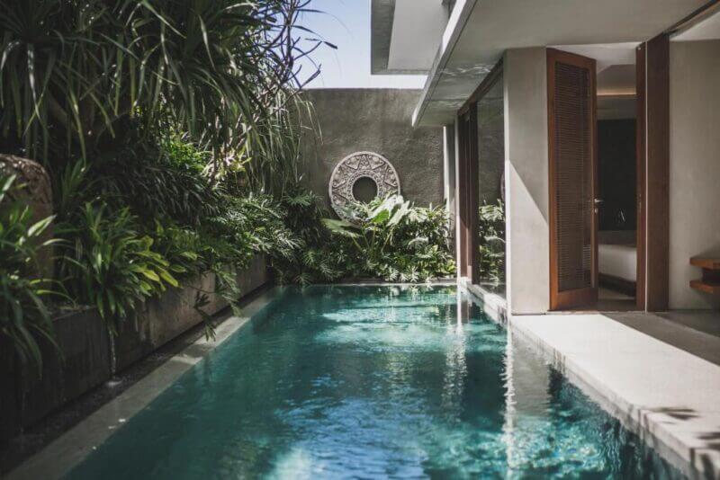 A narrow outdoor swimming pool beside a modern building at Blossom Eco Luxe Villas in Canggu, surrounded by lush green plants and a decorative circular wall feature.