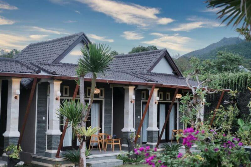 Two single-story Bali Villa units with tiled roofs, front porches, and chairs are surrounded by lush tropical plants and flowers, with a mountain and blue sky in the background—perfect for a peaceful stay at Lava Bali Villa.