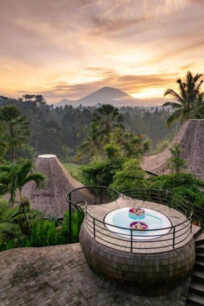 Circular outdoor jacuzzi with floating flowers on a wooden deck at Eco Six Bali overlooks lush tropical trees and distant mountains at sunrise.