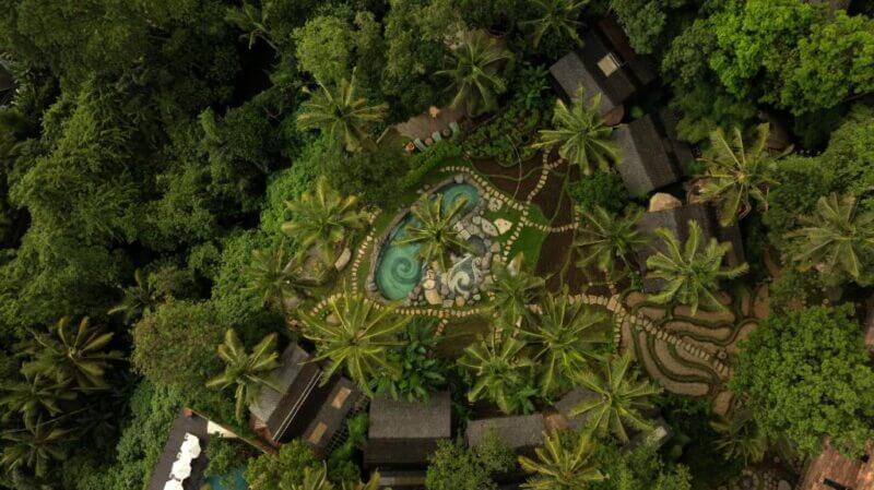 Aerial view of Bambu Indah, an eco resort in Ubud, featuring a central swimming pool surrounded by palm trees, pathways, and several buildings nestled in dense greenery.