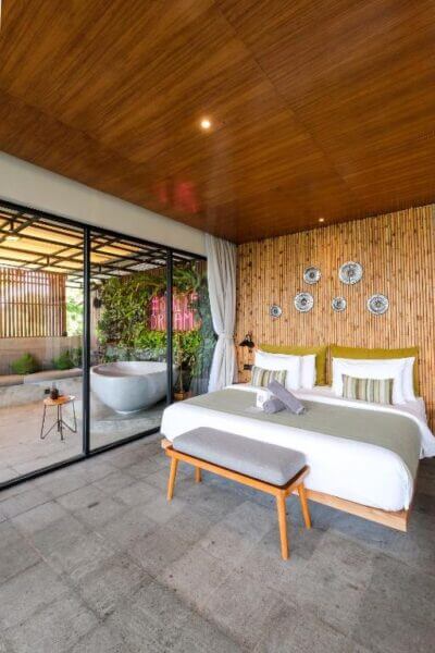 Modern bedroom at Elementum Uluwatu features a wooden ceiling, bamboo accent wall, and large glass doors opening to an outdoor area with a freestanding bathtub and lush greenery—perfect for your ultimate Bali resort escape.