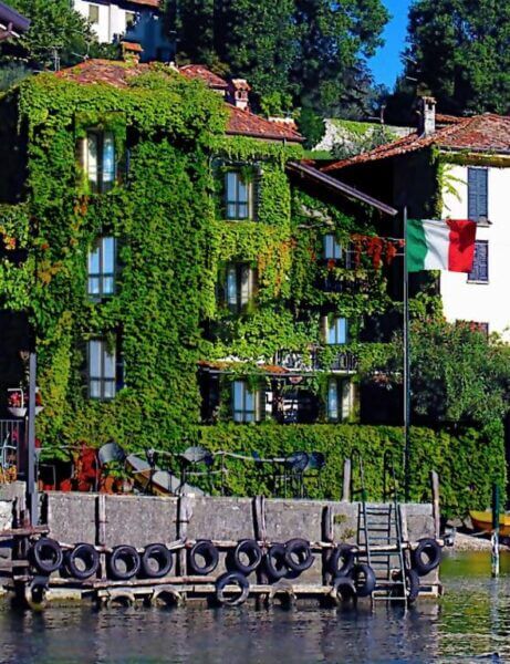 A building covered in green ivy stands by the Bellagio waterfront with a dock, outdoor seating, and an Italian flag—perfectly capturing the charm of Pescallo Apartments.