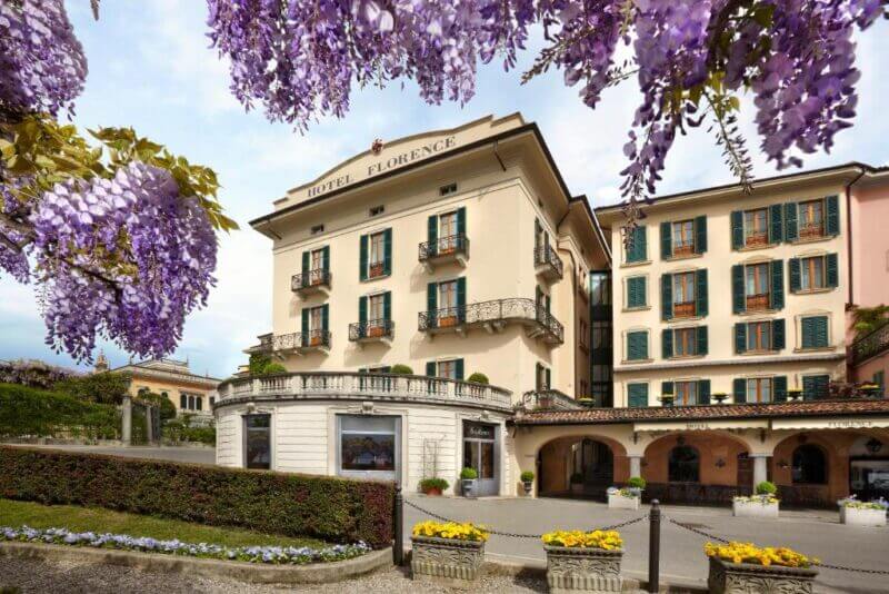 A three-story building labeled Hotel Florence, reminiscent of Bellagio elegance, features balconies and is surrounded by purple flowering trees and yellow flowers in planters.