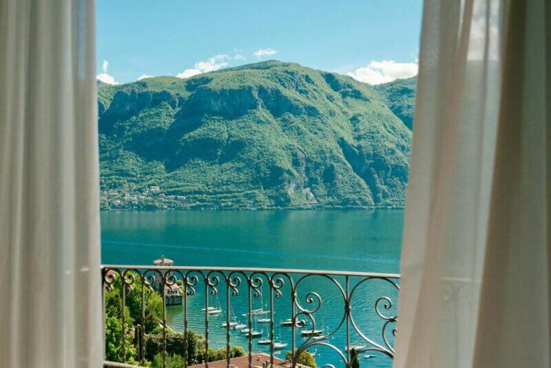 A view of a lake and green mountains seen through open white curtains and an ornate balcony railing at Hotel Belvedere Bellagio, with boats drifting on the water below.