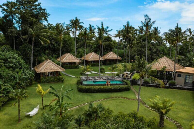 Aerial view of Lempuyang Hotel, a tropical resort in Ujung with thatched-roof villas, a central swimming pool, palm trees, and landscaped green lawns under a blue sky.