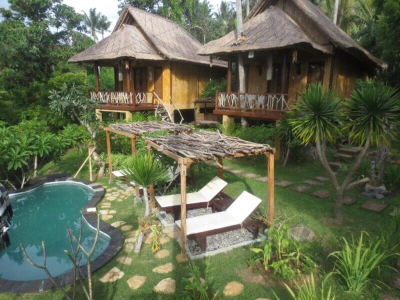 Two wooden cottages with thatched roofs at Jepun Didulu Cottages overlook a small, curved swimming pool and lounge chairs in a lush, tropical garden setting near Ujung.