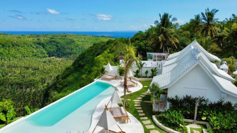 Atalaya Villas in Nusa Penida offers a stunning white villa with an infinity pool overlooking a lush green valley and distant ocean, all beneath a clear blue sky, surrounded by tropical plants and palm trees.