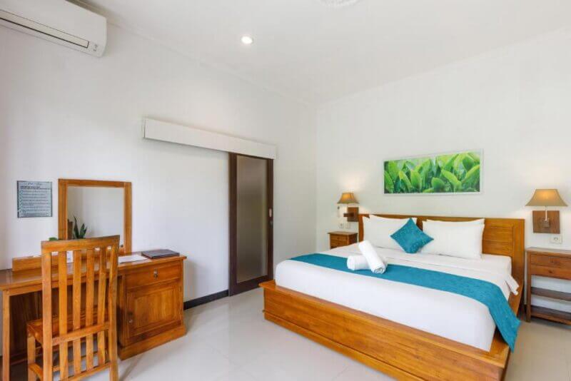 A tidy hotel room at Ped Cottage Nusa Penida features a wooden double bed, a desk and chair, white walls, air conditioner, and green artwork above the bed. Fresh towels are placed neatly on the bed.