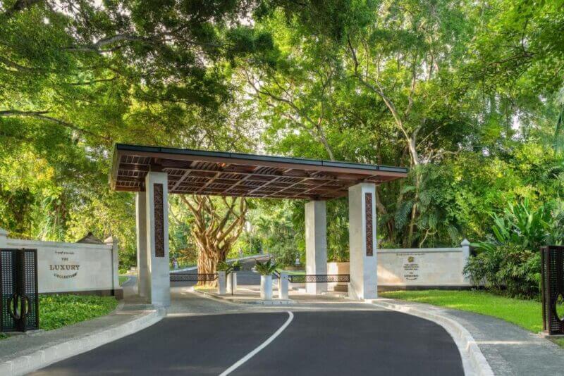 Entrance gate with a modern canopy and white pillars, surrounded by lush green trees and landscaped gardens, welcomes you into Laguna Nusa Dua, a Bali luxury hotel with a stunning driveway leading through.